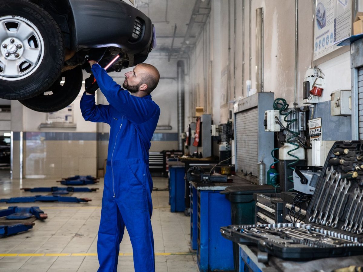 Kfz-Mechaniker im Blaumann prüft die Unterseite eines Fahrzeugs auf der Hebebühne