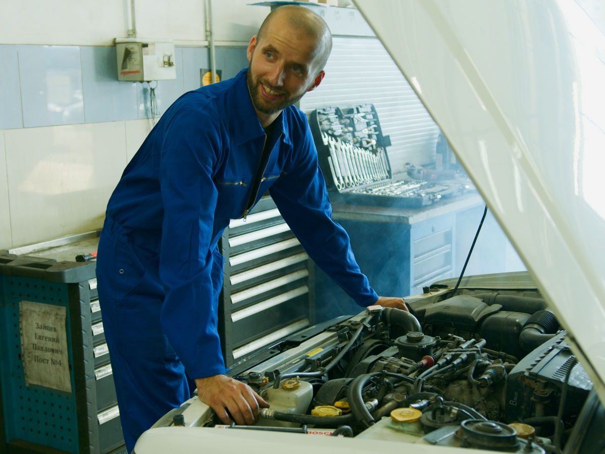Lächelnder Kfz-Mechaniker im Blaumann arbeitet am Motor unter der geöffneten Motorhaube